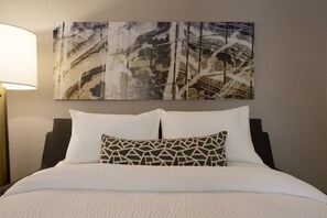 Un lit bien fait, avec une literie blanche et un coussin à motifs géométriques, un tableau abstrait accroché au mur et une lampe sur pied placée à gauche.