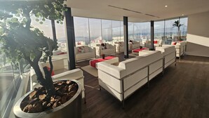 A modern lounge area with white leather sofas, a large potted plant, and a view of the cityscape through large windows.