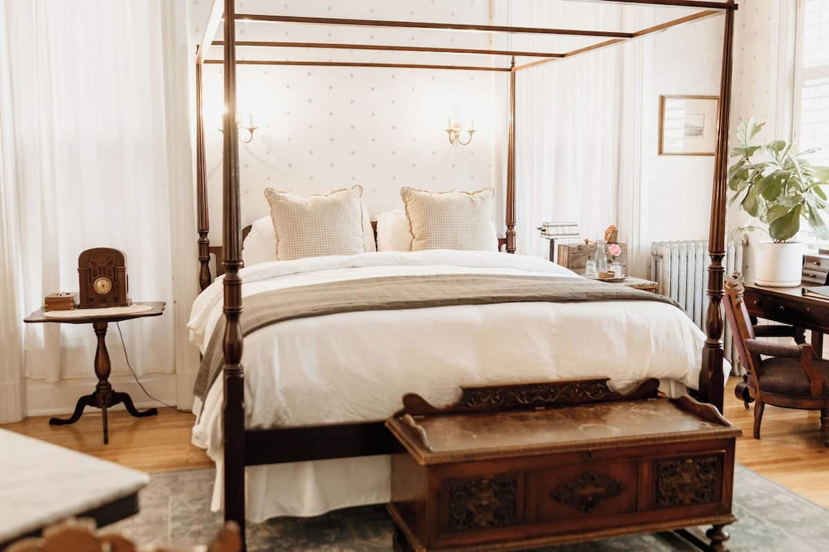 Una cama con dosel y tablero de madera, una mesita de noche con una radio, y una planta grande en maceta.