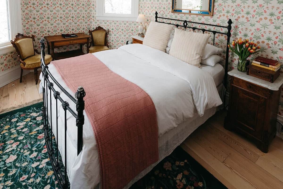 Un dormitorio con cama, mesita de noche, espejo, un cuadro en la pared y papel tapiz estampado con motivos florales.