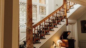 Una imponente escalera con balaustres de madera y un columnario de mármol, que conduce a una sala con un piano y un radiador.