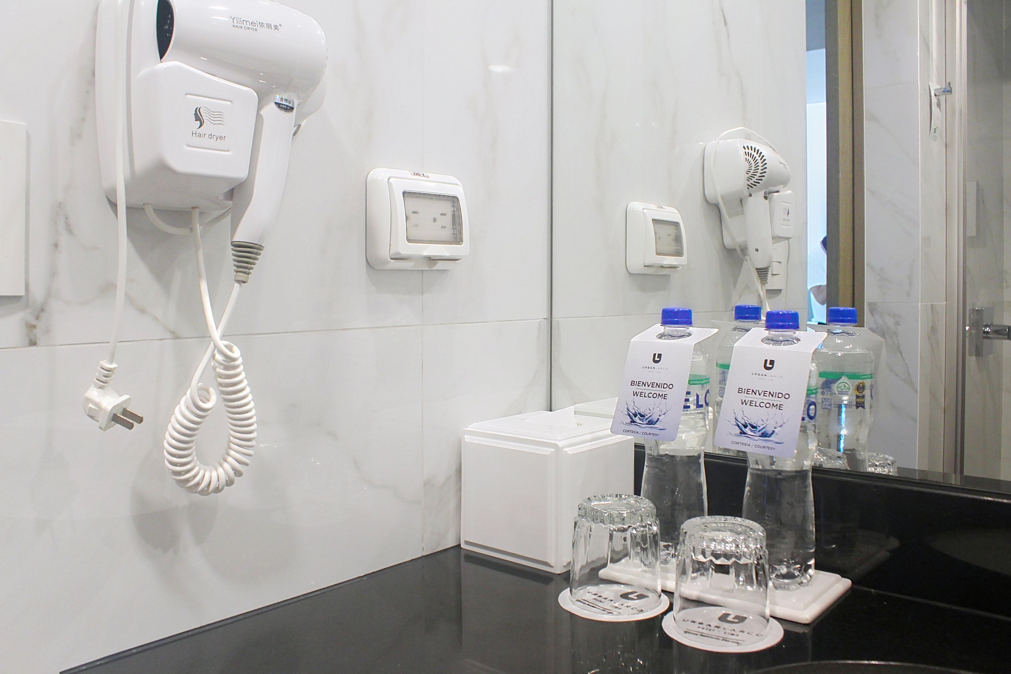 Un baño de hotel con secador de pelo, una caja blanca pequeña y botellas de agua con etiquetas que dicen 'BIENVENIDO WELCOME'.