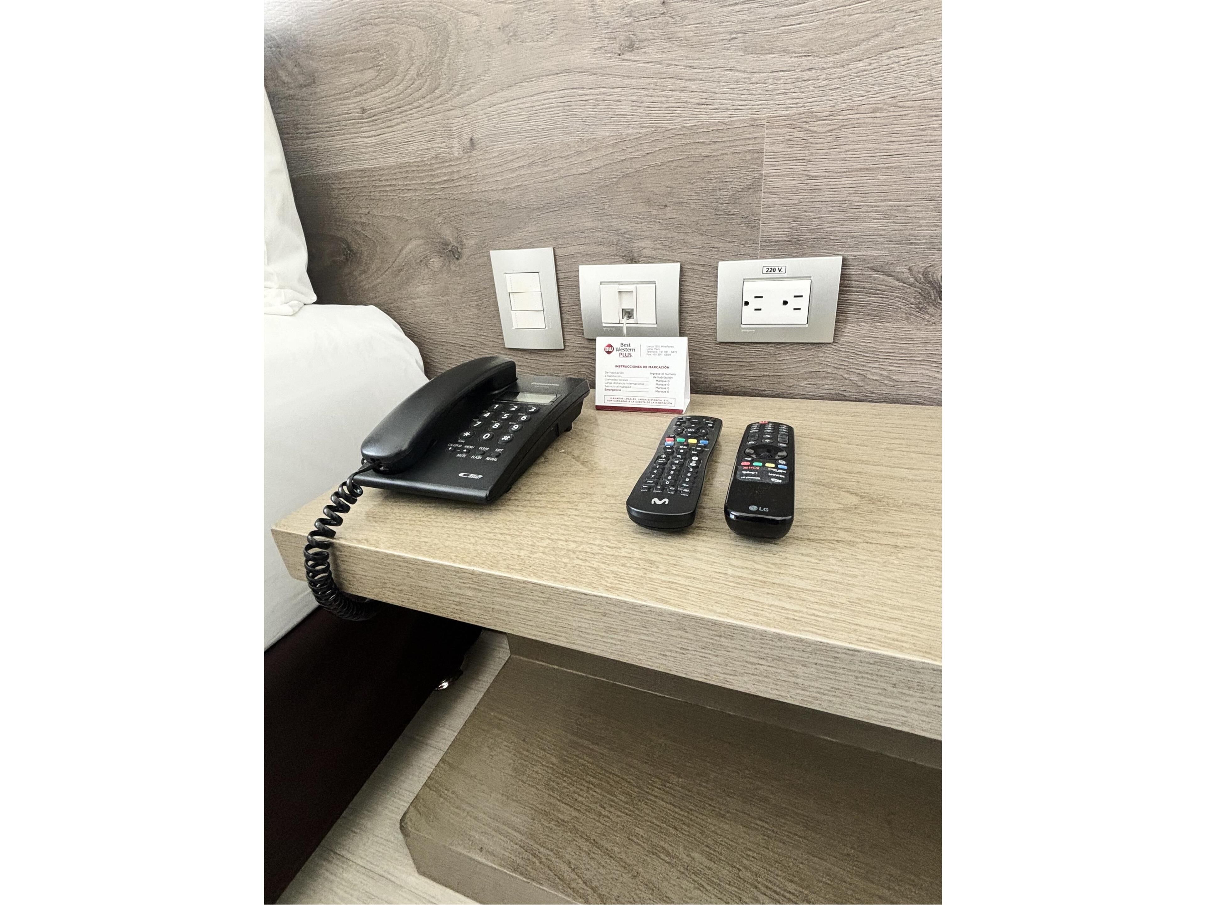 A hotel room with a wooden headboard, a telephone, and two remote controls on a wooden table.