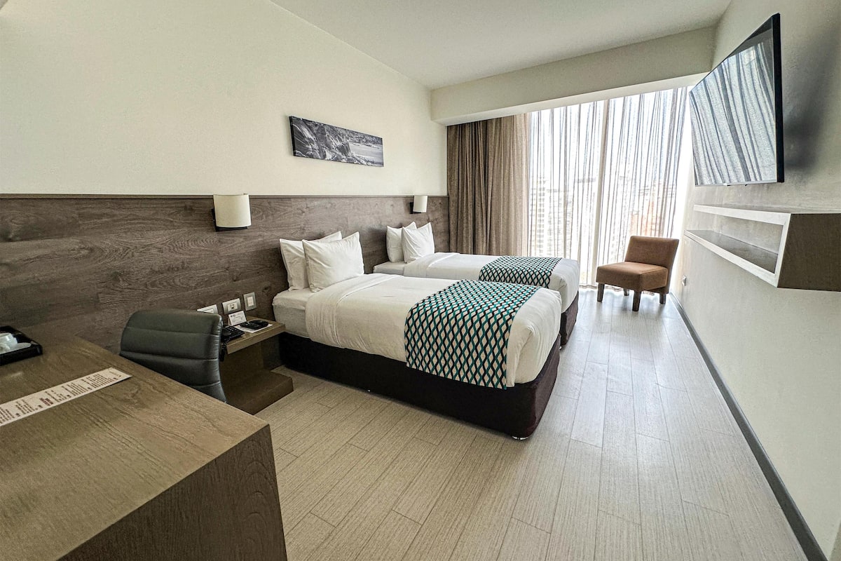 Quarto de hotel com duas camas, uma mesa, uma cadeira, uma televisão e uma janela com cortinas.