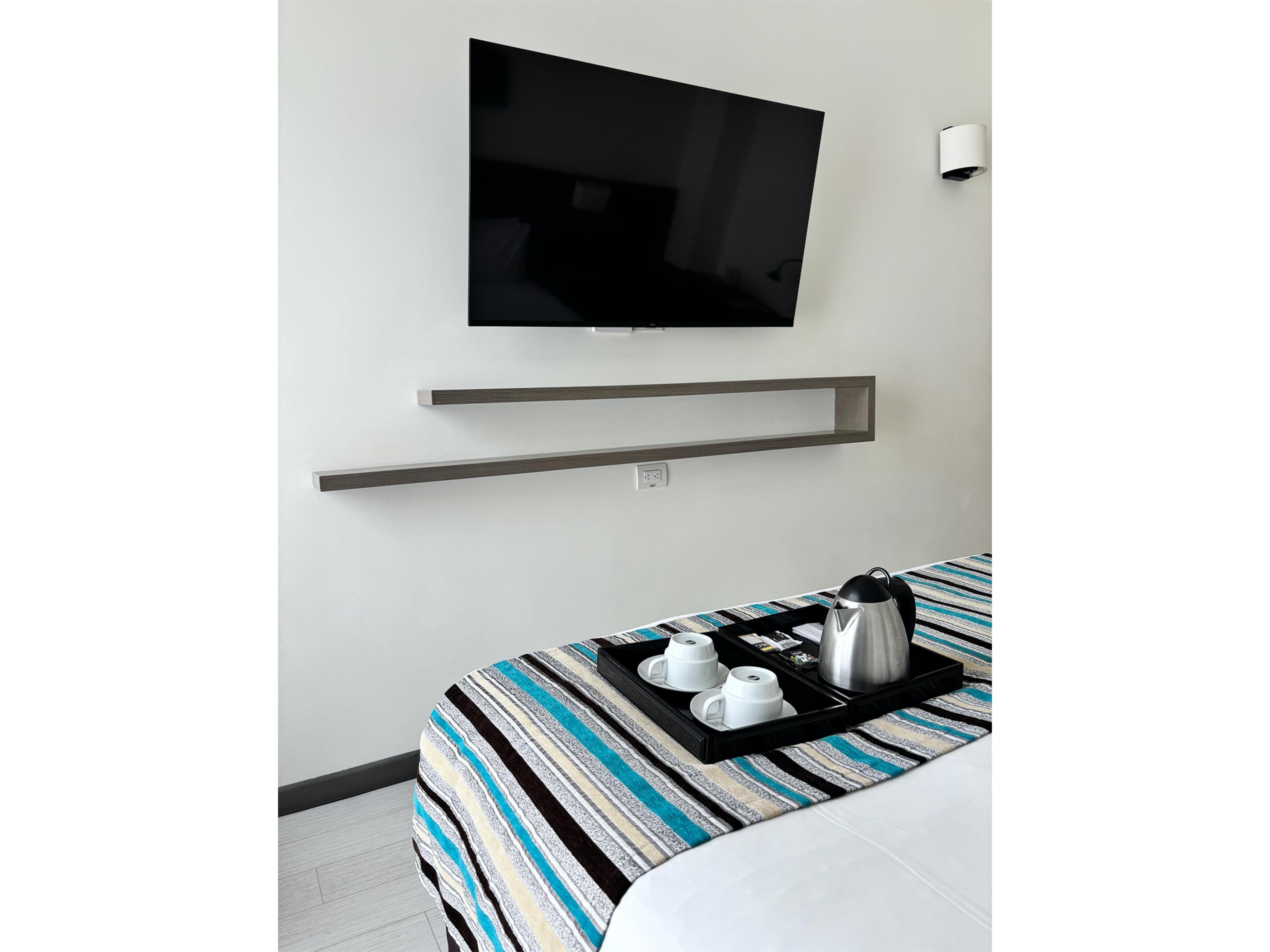 Une chambre d’hôtel avec un lit, un téléviseur fixé au mur, une étagère au-dessus du lit et un plateau avec des tasses et une bouilloire posé sur le lit.