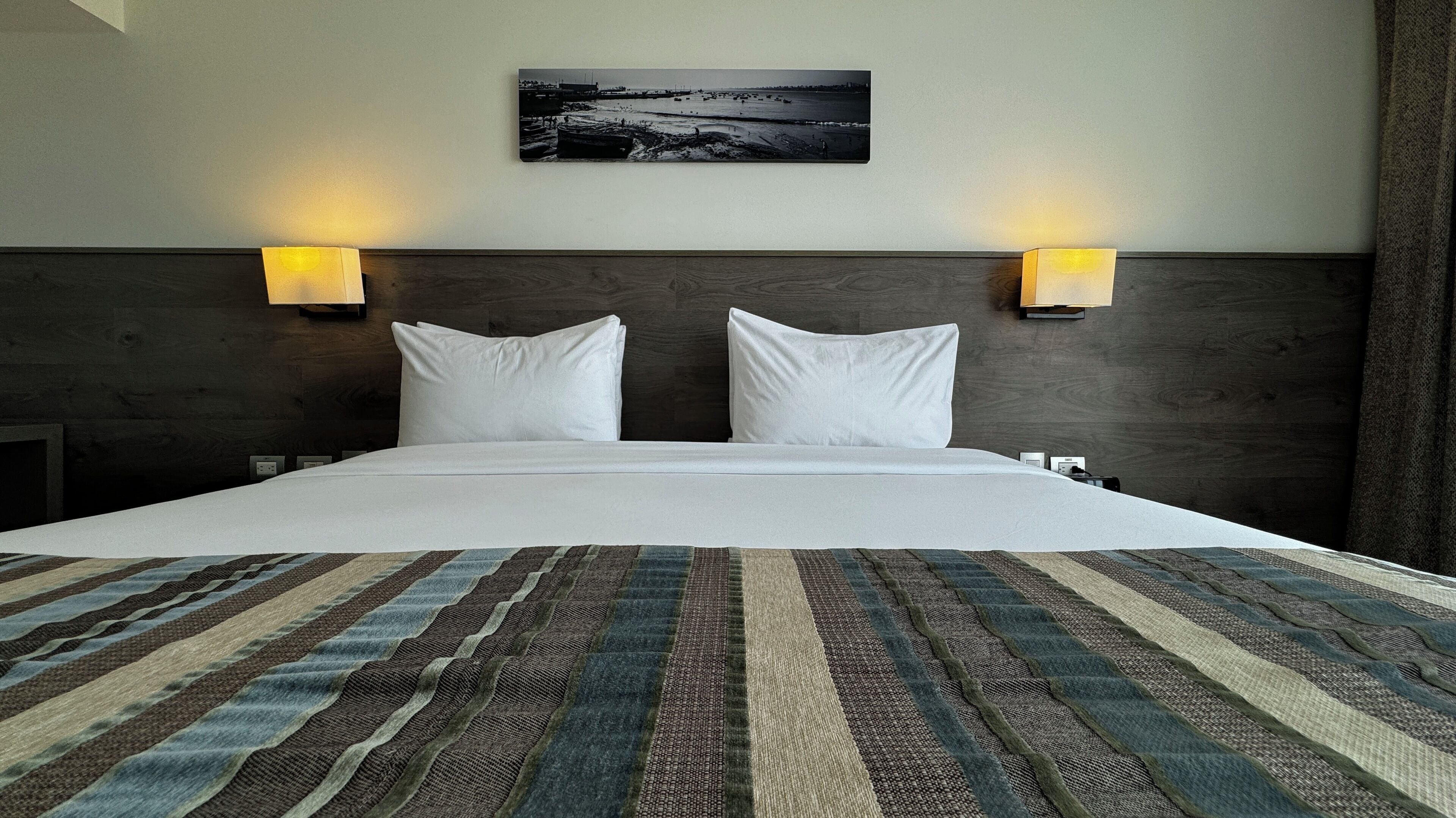 A hotel room with a large bed, two wall-mounted lamps, a headboard with a dark wood finish, and a black and white photograph on the wall.