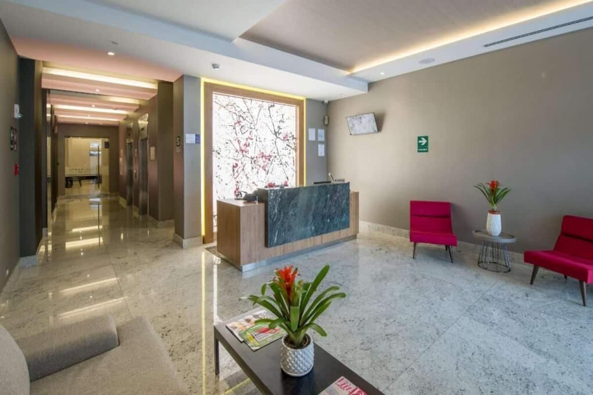 Um lobby de hotel com balcão de recepção, área de espera com cadeiras vermelhas, balcão de atendimento e um corredor que dá acesso a outros quartos.