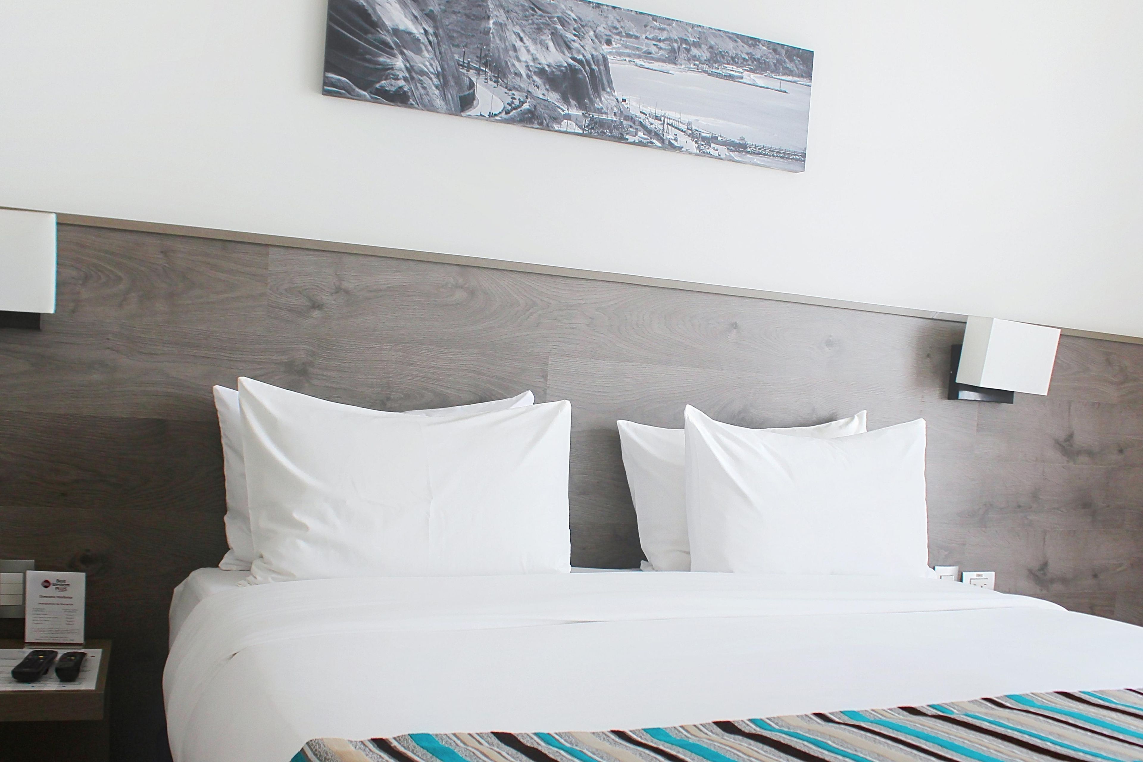 Una habitación de hotel con cama, dos almohadas, cabecera, mesita de noche con lámpara y un cuadro en la pared.
