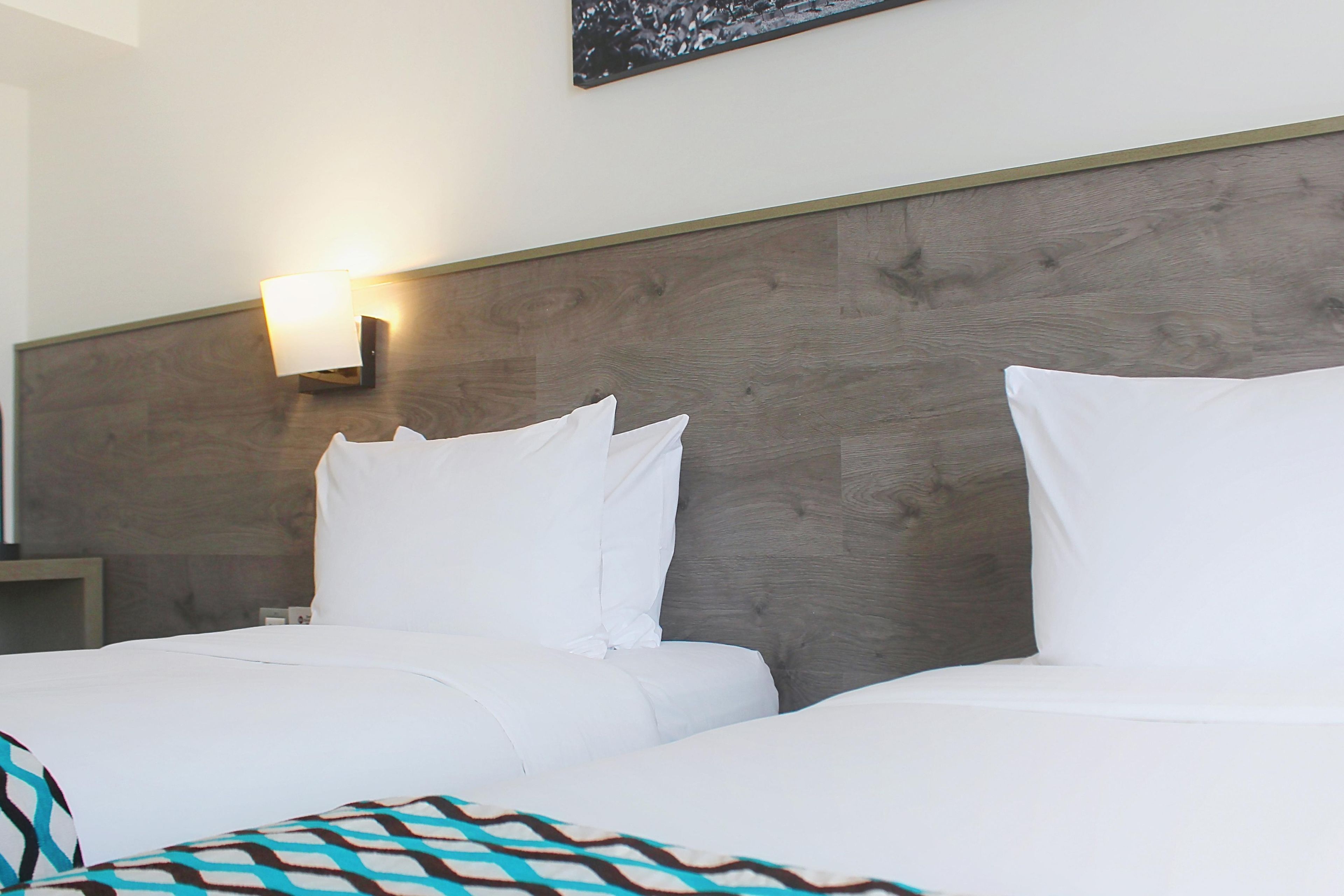 A hotel room with a wooden headboard, two beds with white bedding, a nightstand, and a wall-mounted lamp.