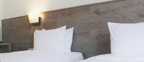 A hotel room with a wooden headboard, two beds with white bedding, a nightstand, and a wall-mounted lamp.