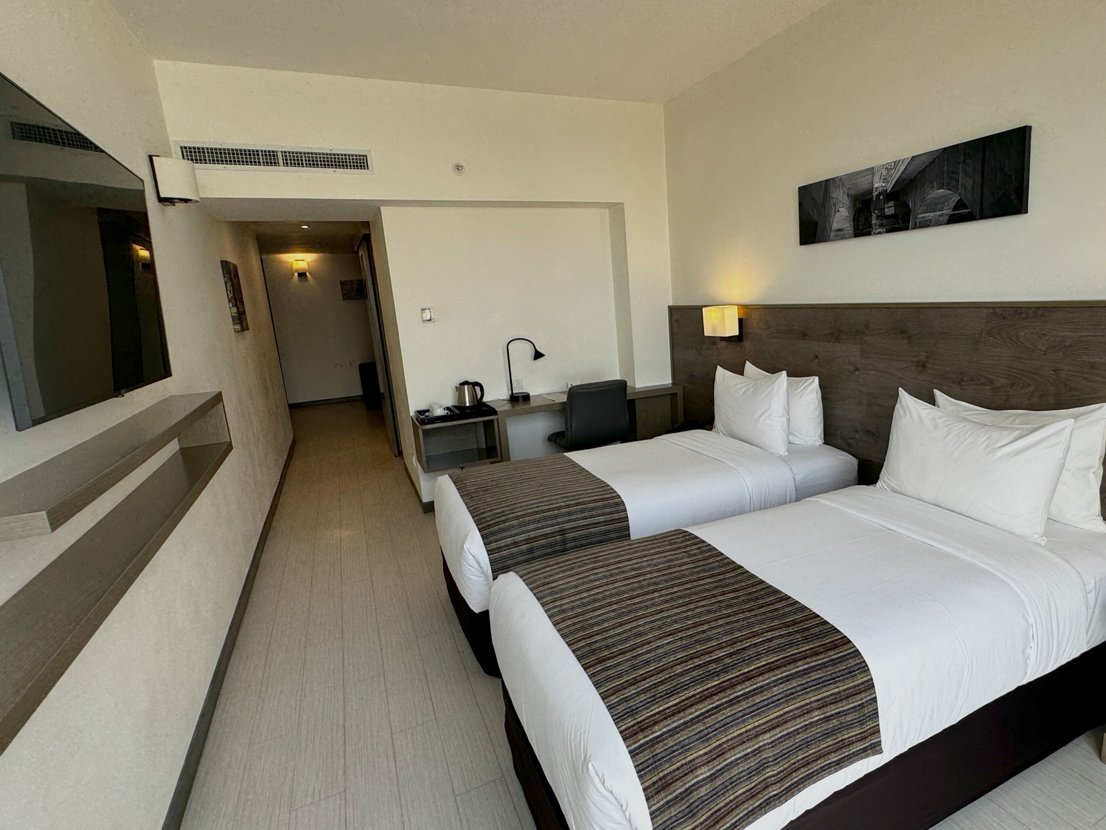 Habitación de hotel con dos camas, un escritorio con silla, un televisor y un cuadro en la pared.