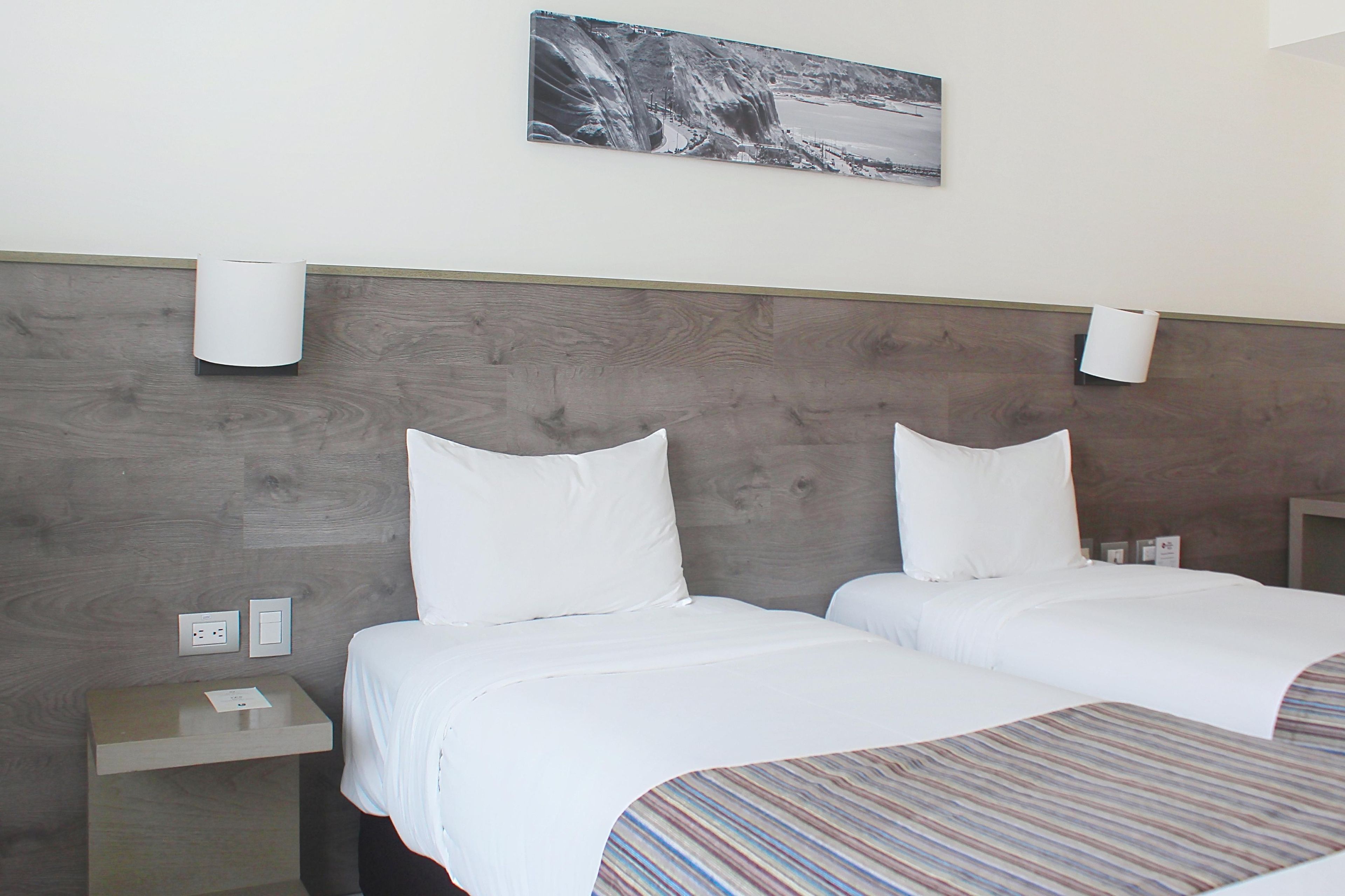 A hotel room with two beds, a wooden headboard, a nightstand, and a wall-mounted light fixture.
