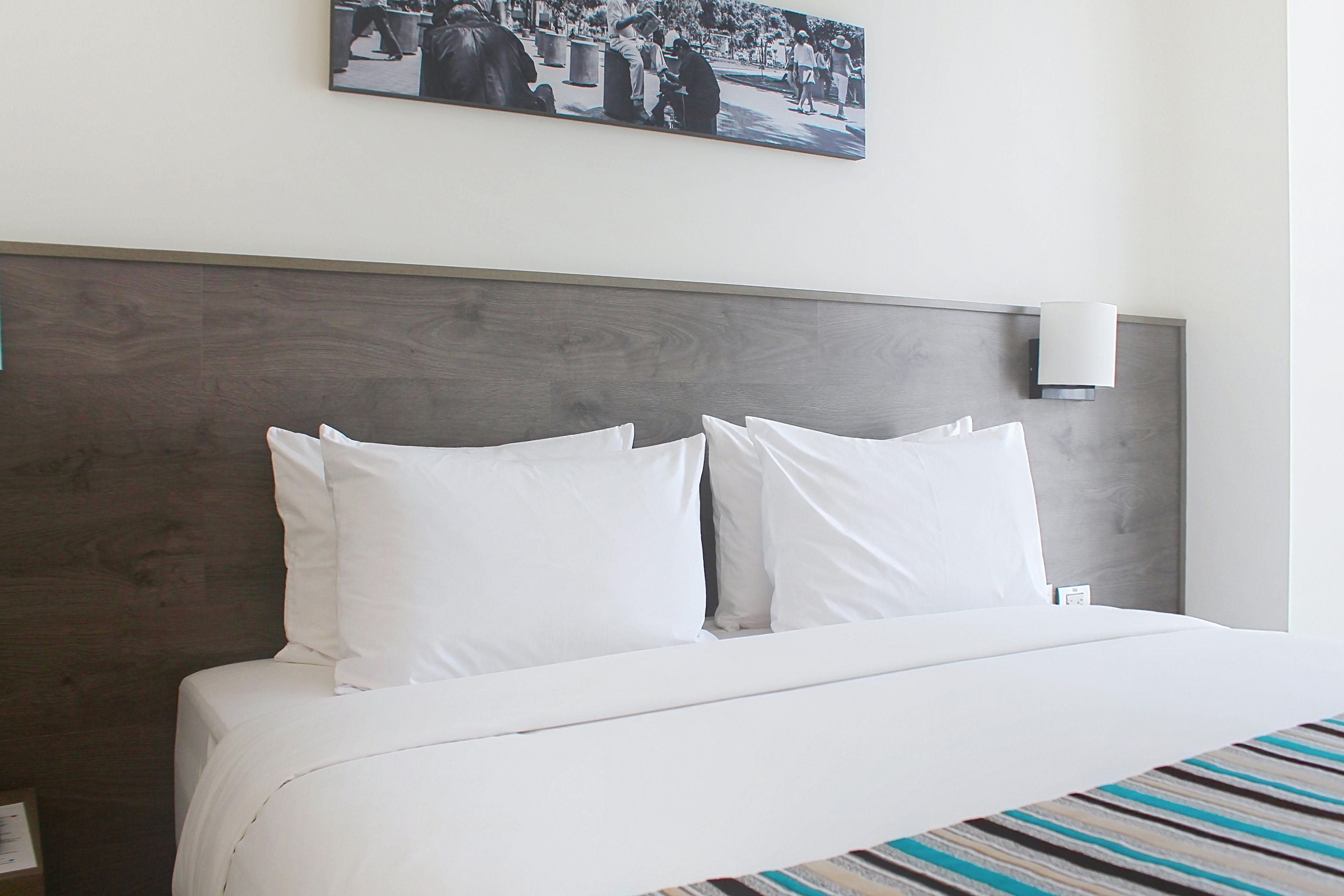 A hotel room with a bed, white bedding, a headboard, a wall-mounted light, and a framed picture above the bed.