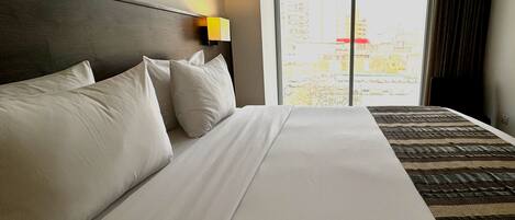 Habitación de hotel con una cama grande, cabecera, mesita de noche con lámpara y vista a edificios a través de la ventana.