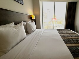 Habitación de hotel con una cama grande, cabecera, mesita de noche con lámpara y vista a edificios a través de la ventana.