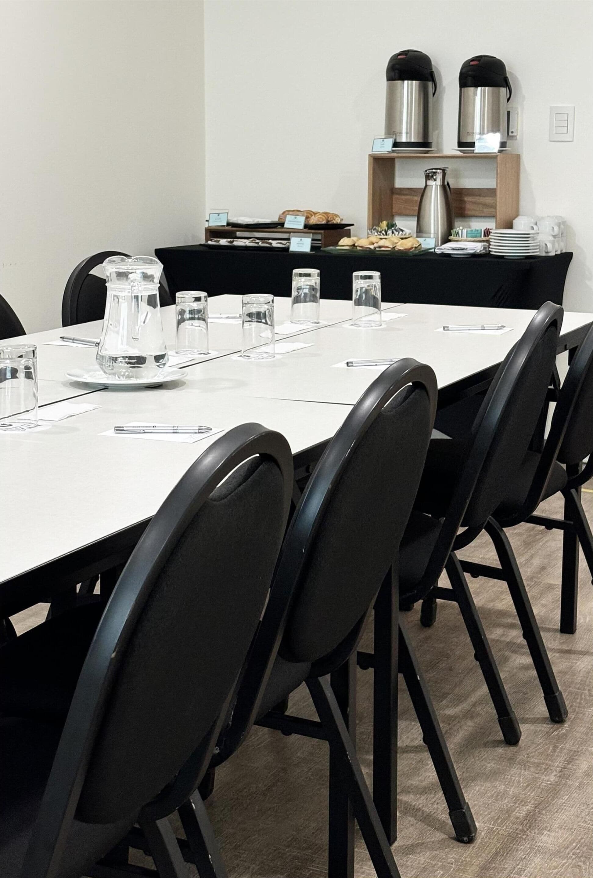 A conference room with a long table, black chairs, and a coffee station with two coffee makers.