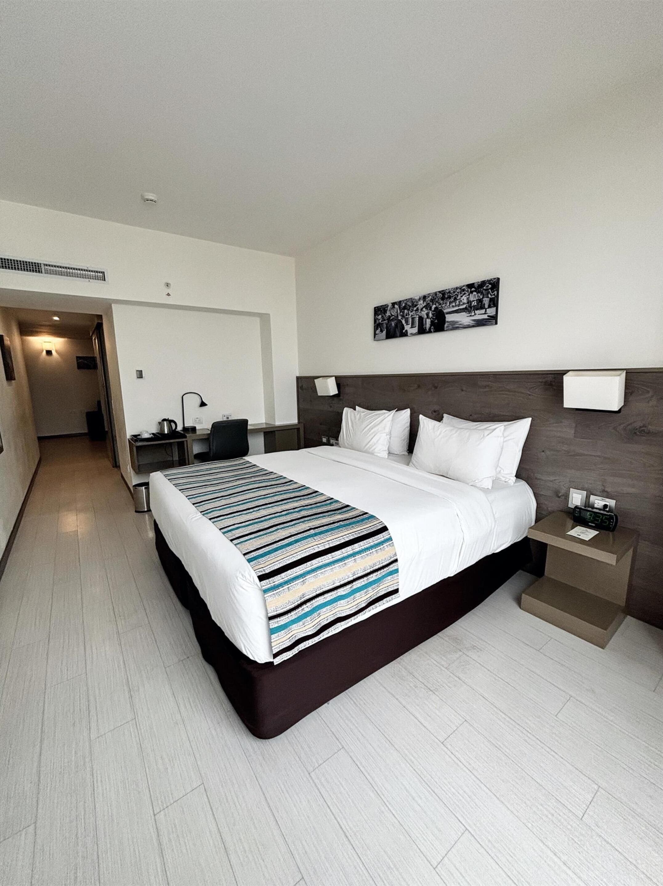 Una habitación de hotel moderna con una cama grande, una mesita de noche con un reloj, un escritorio con una silla y un pasillo que conduce a otra habitación.