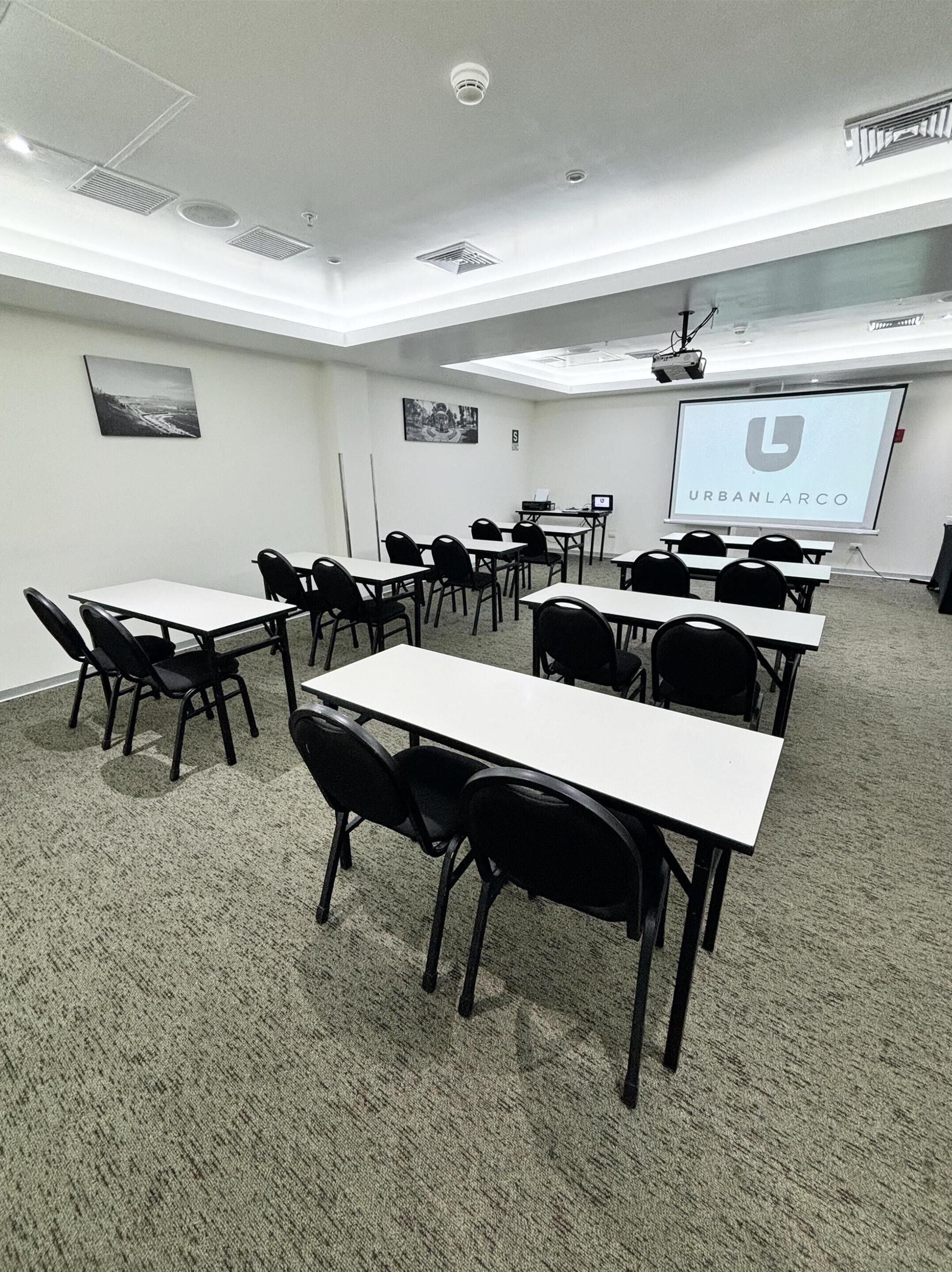 A conference room with white tables and black chairs, a projector screen displaying 'URBANLARCO', and a ceiling with recessed lighting and ventilation.