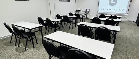 A conference room with white tables and black chairs, a projector screen displaying 'URBANLARCO', and a ceiling with recessed lighting and ventilation.