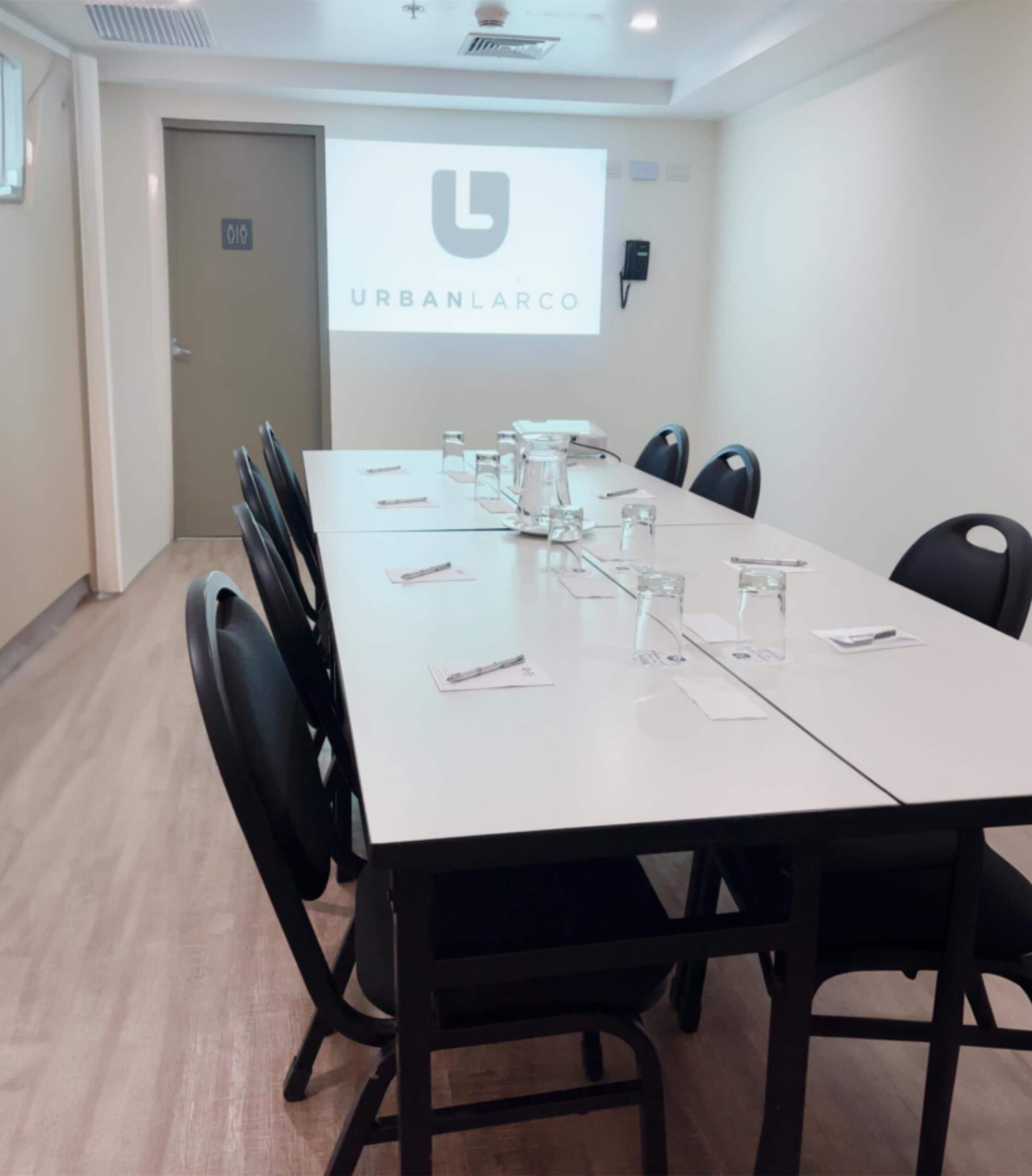 A conference room with a long table, chairs, and a projector screen displaying "URBANLARCO".