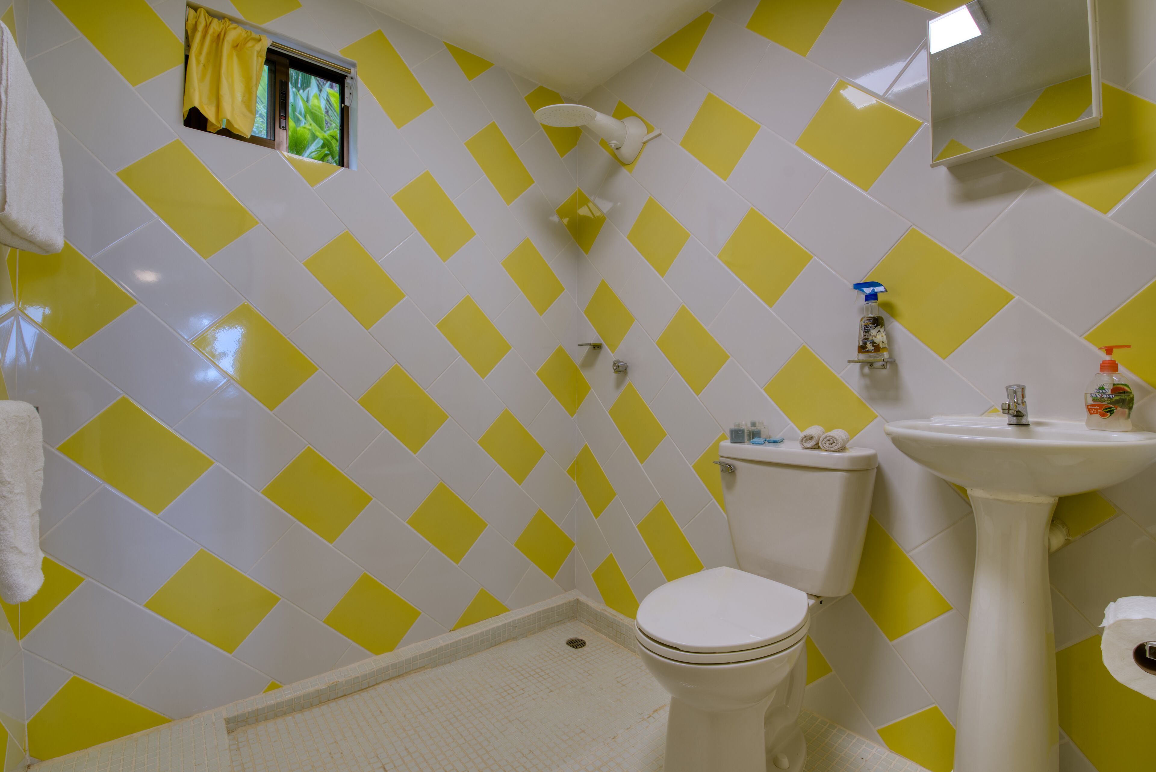 Une salle de bain avec un mur recouvert de carreaux blancs et jaunes formant un motif losangique, des toilettes blanches, un lavabo sur pied et une fenêtre avec un rideau jaune.
