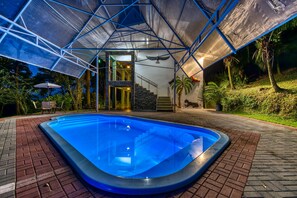 Una piscina con una estructura de techo geométrica, rodeada por un camino de ladrillo y plantas tropicales.
