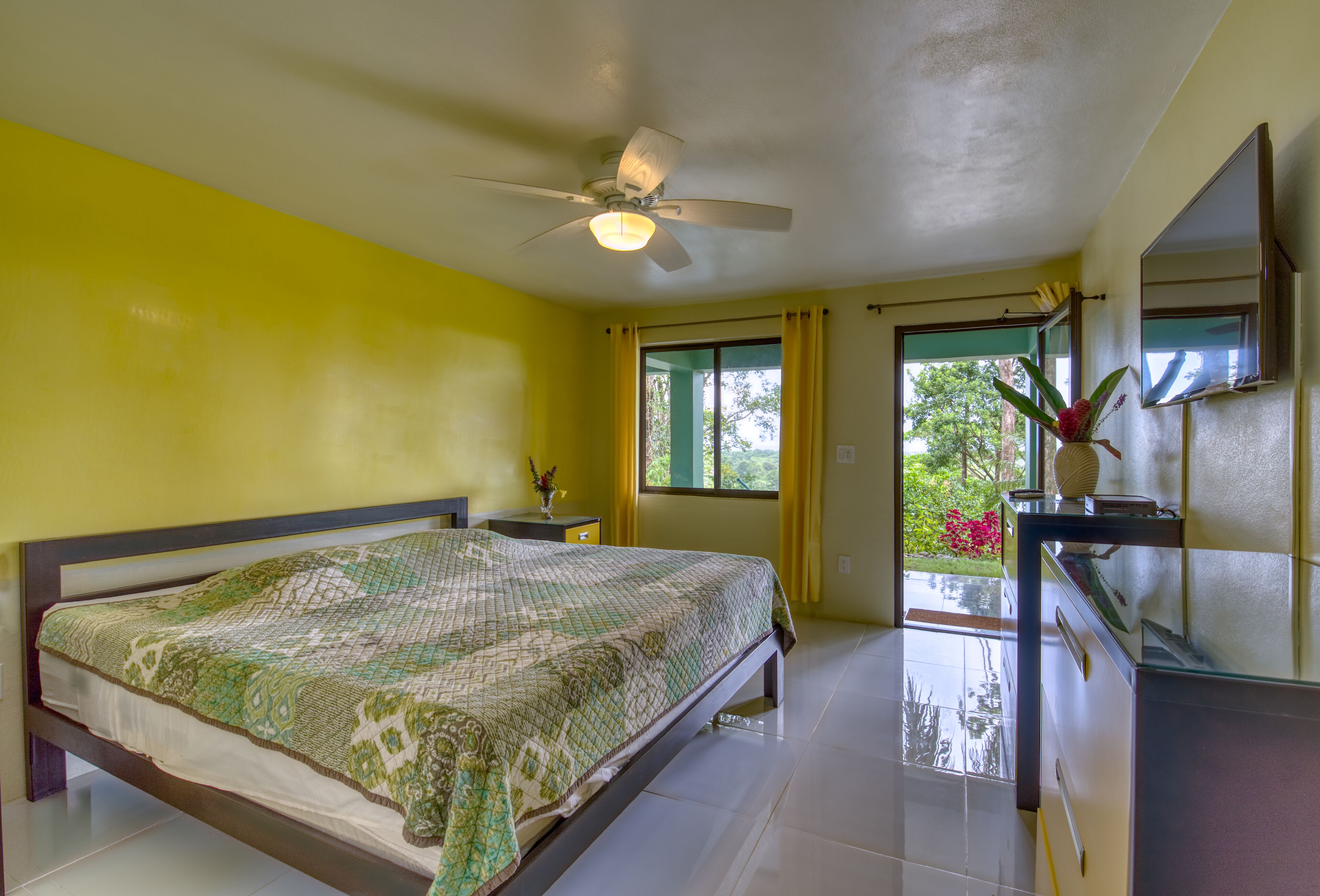 A bedroom with a bed, a ceiling fan, a nightstand, a dresser, a mirror, and a window with curtains.