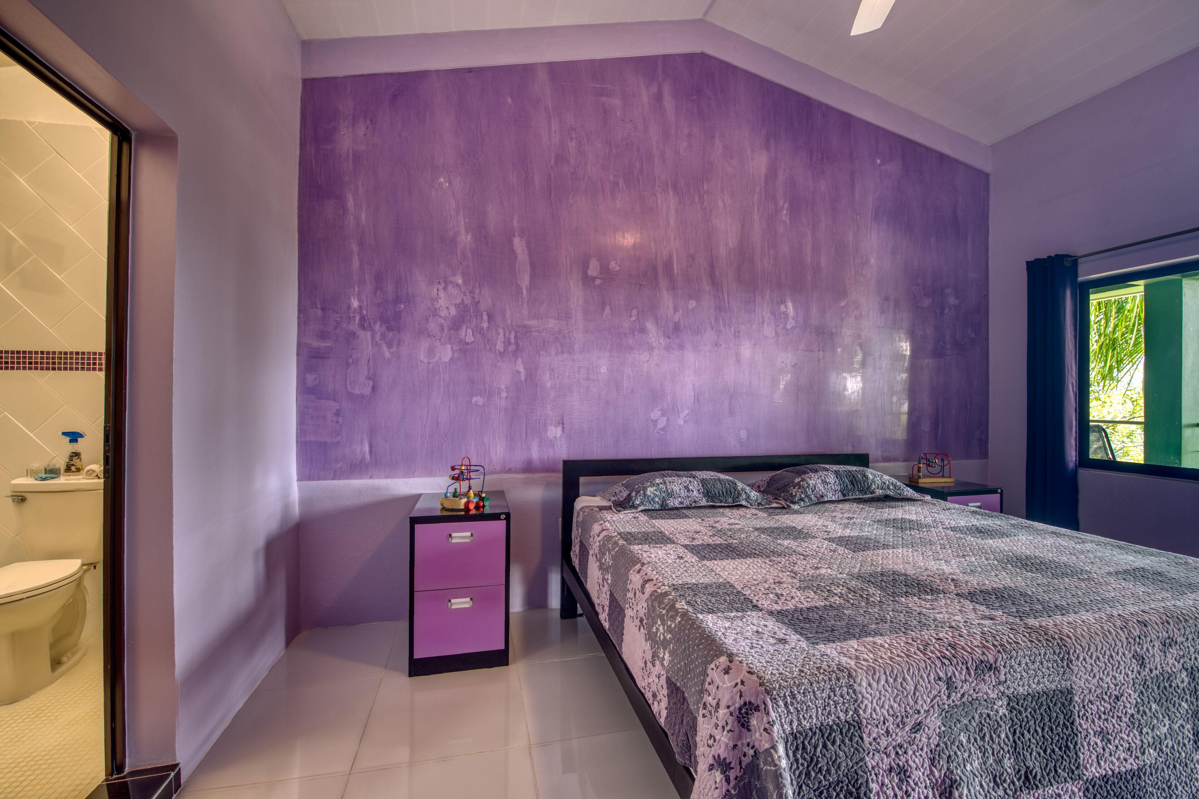 Un dormitorio con una pared de acento color púrpura, una cama con colcha estampada, una mesita de noche con lámpara y una ventana con vistas a la vegetación.