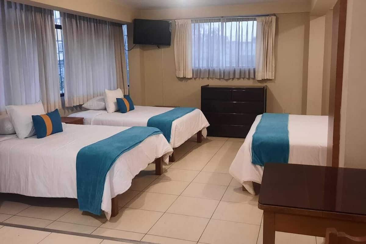 Habitación de hotel con tres camas, un televisor montado en la pared, una mesa de madera y una ventana con cortinas.