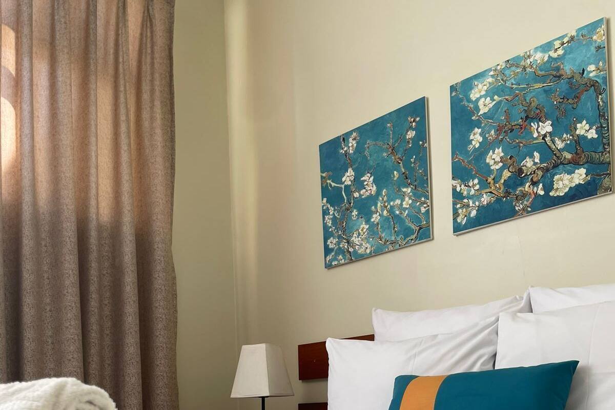 Habitación de hotel con una cama bien hecha, una pila de toallas, una lámpara y dos cuadros con motivos florales en la pared.