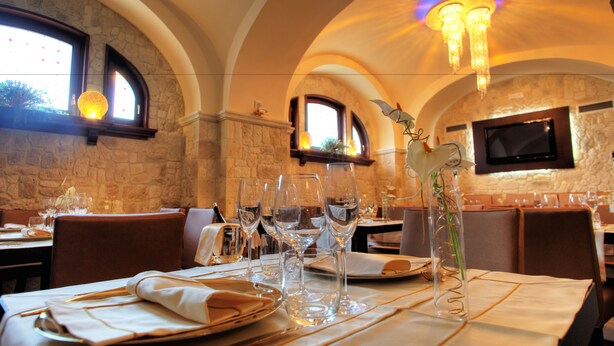 Un restaurante con paredes de piedra, ventanas arqueadas y una araña. Las mesas están dispuestas con copas de vino, platos y cubiertos.