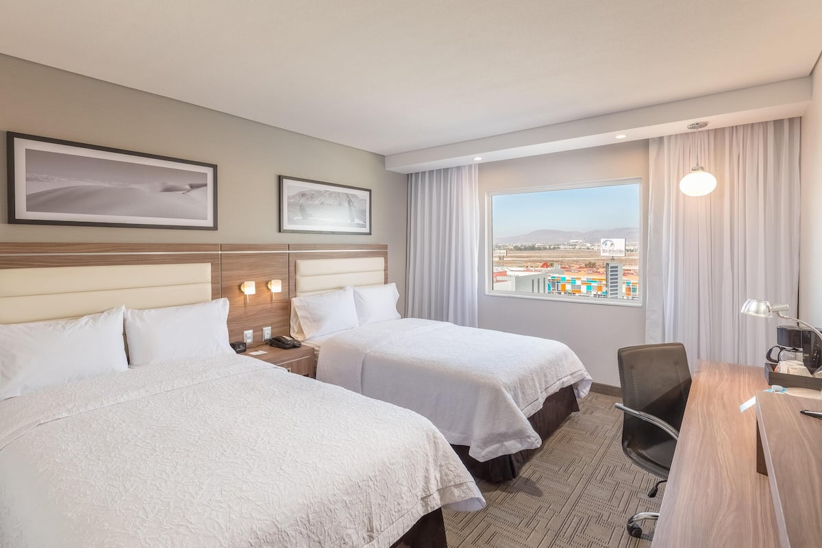 A hotel room with two beds, a desk, a chair, a nightstand, and a window with a city view.