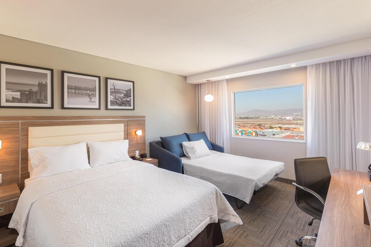 A hotel room with two beds, a desk, a chair, a view of the city, and framed pictures on the wall.