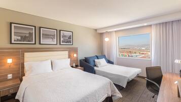 A hotel room with two beds, a desk, a chair, a view of the city, and framed pictures on the wall.