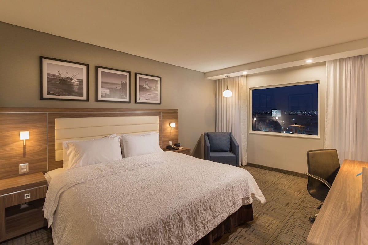 A hotel room with a bed, two bedside tables, a desk, a chair, a window with a city view, and three framed pictures on the wall.