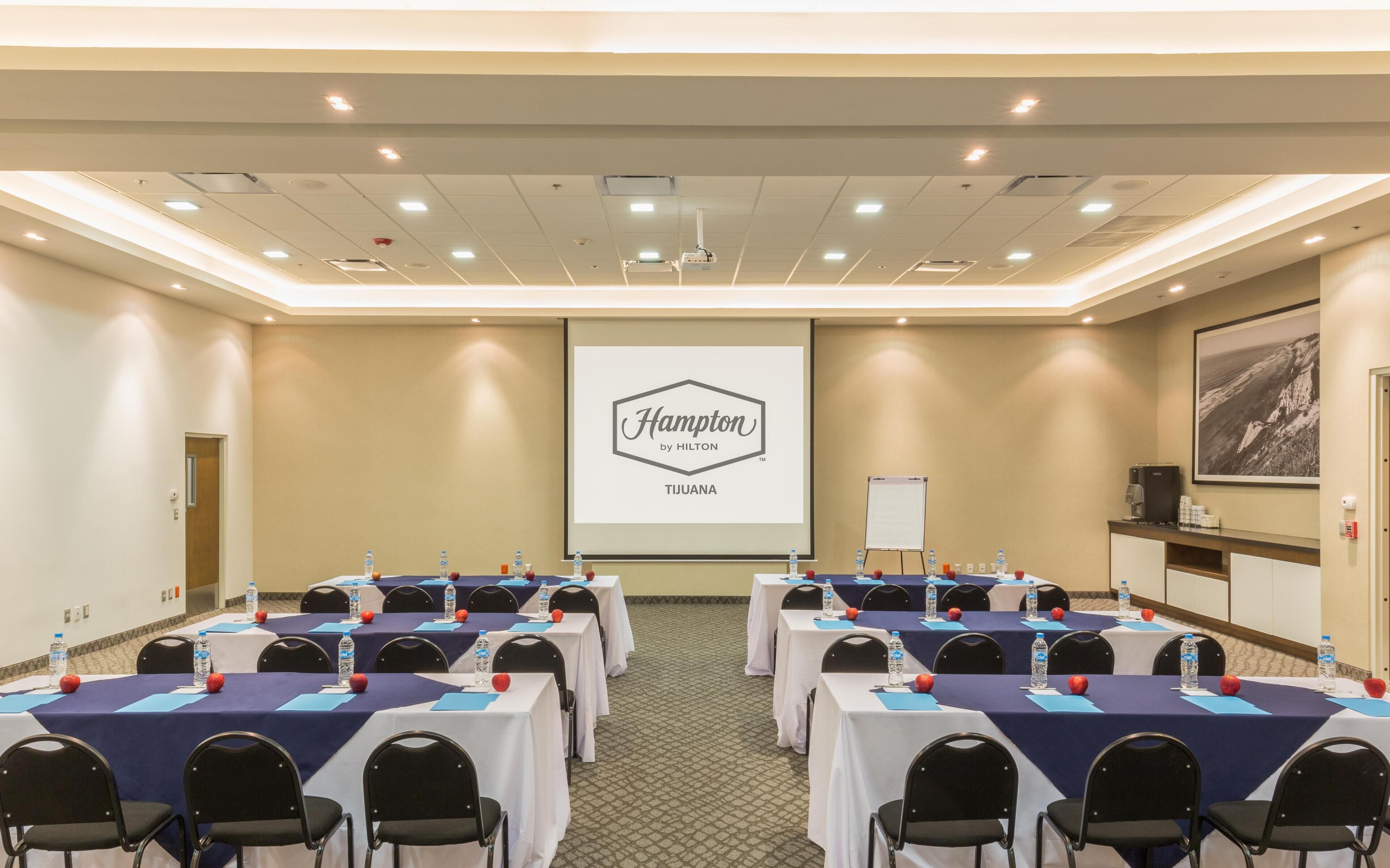 A conference room with multiple tables, chairs, a projector screen, and a presentation on the wall.
