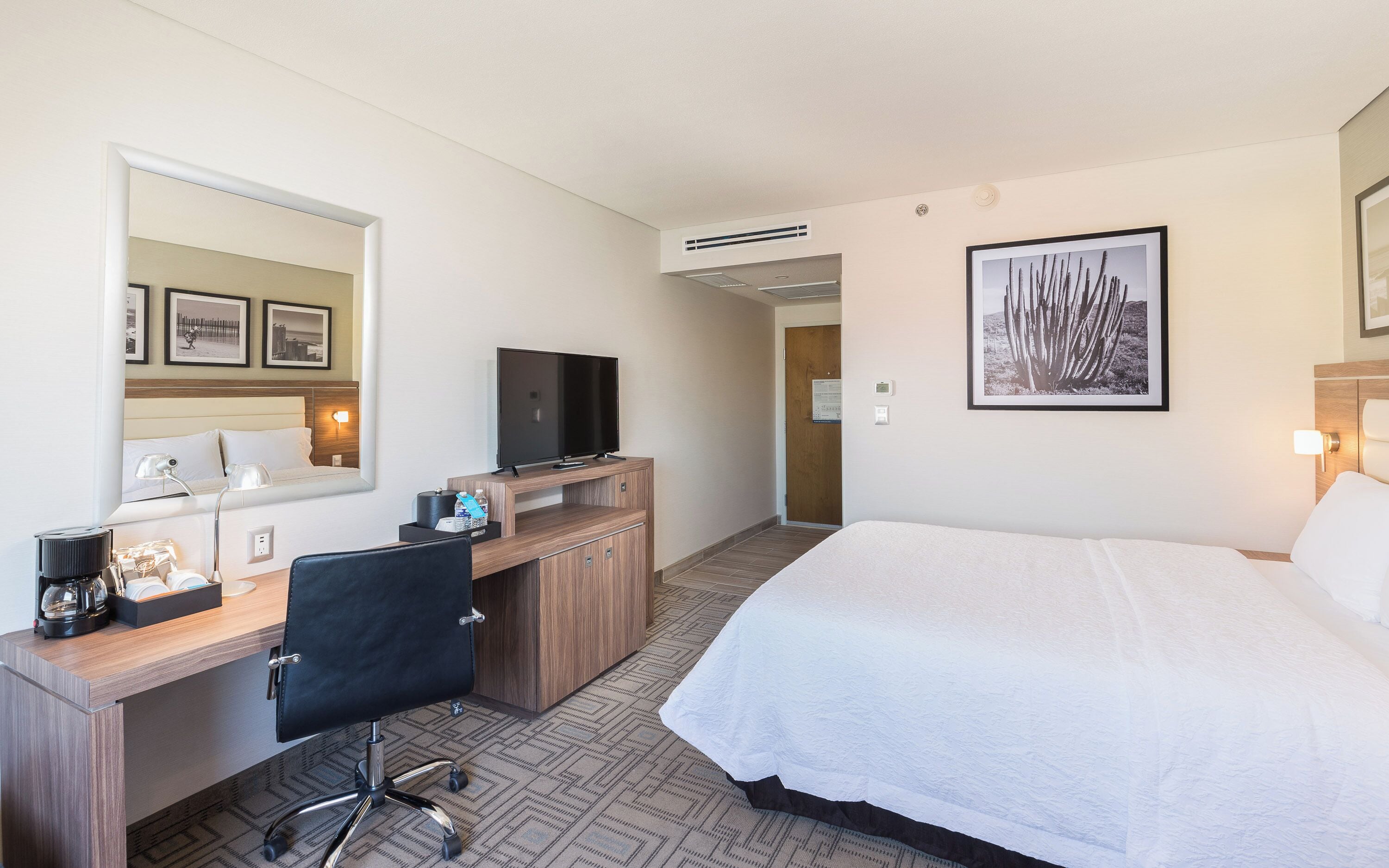 Una habitación de hotel con una cama grande, un escritorio con televisor, una silla, un espejo y un cuadro en la pared.