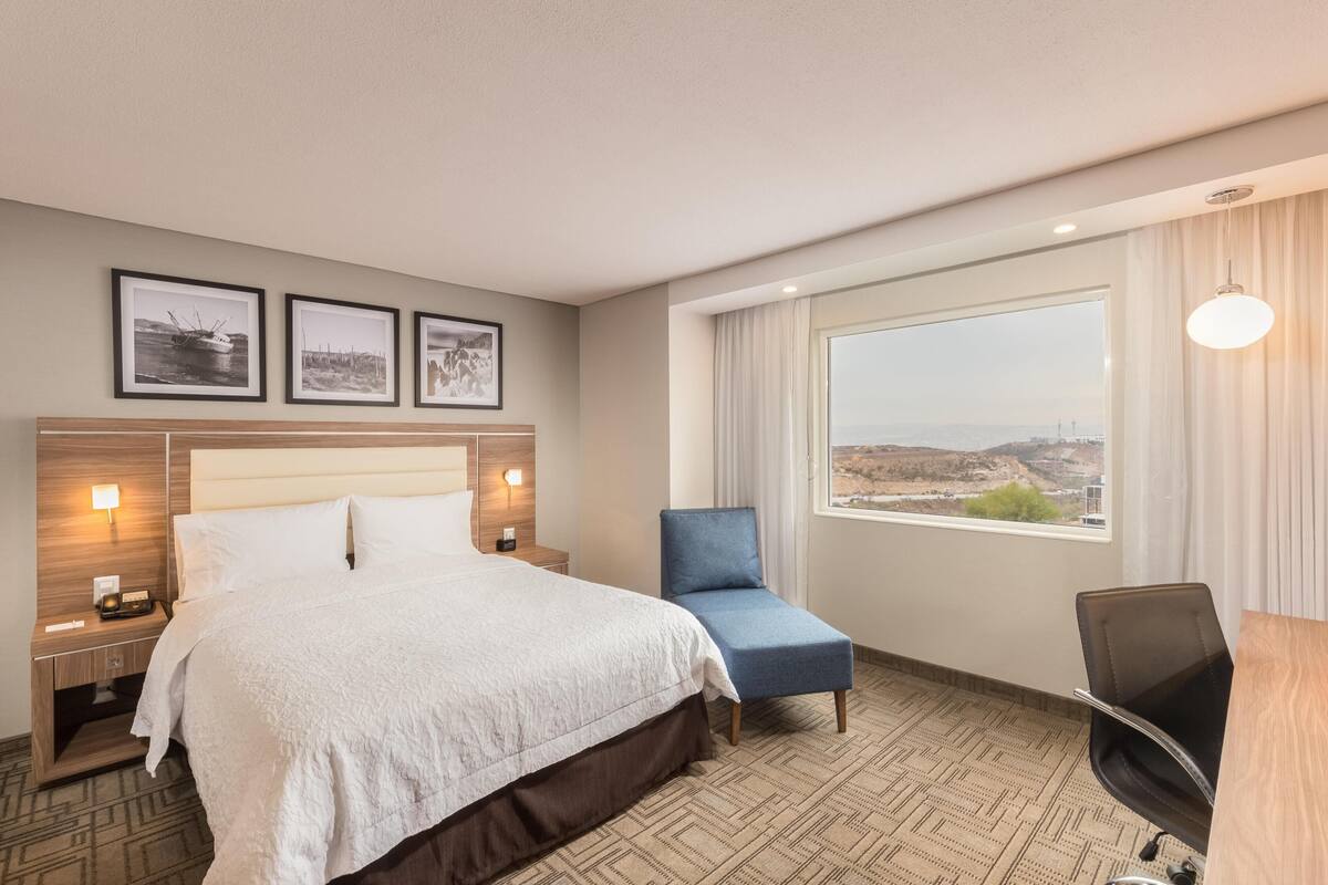 A hotel room with a bed, a desk, a chair, a large window with a view, and three framed pictures on the wall.