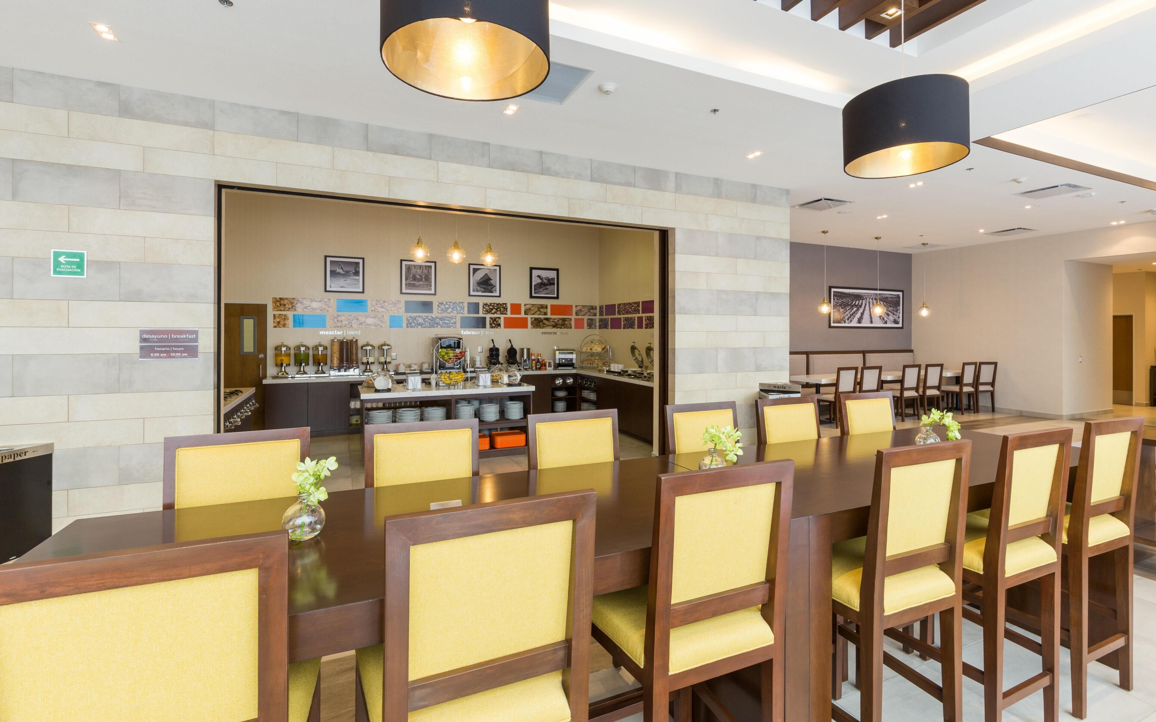 A hotel dining area with a long table, chairs, a bar, and a tiled wall with artwork.