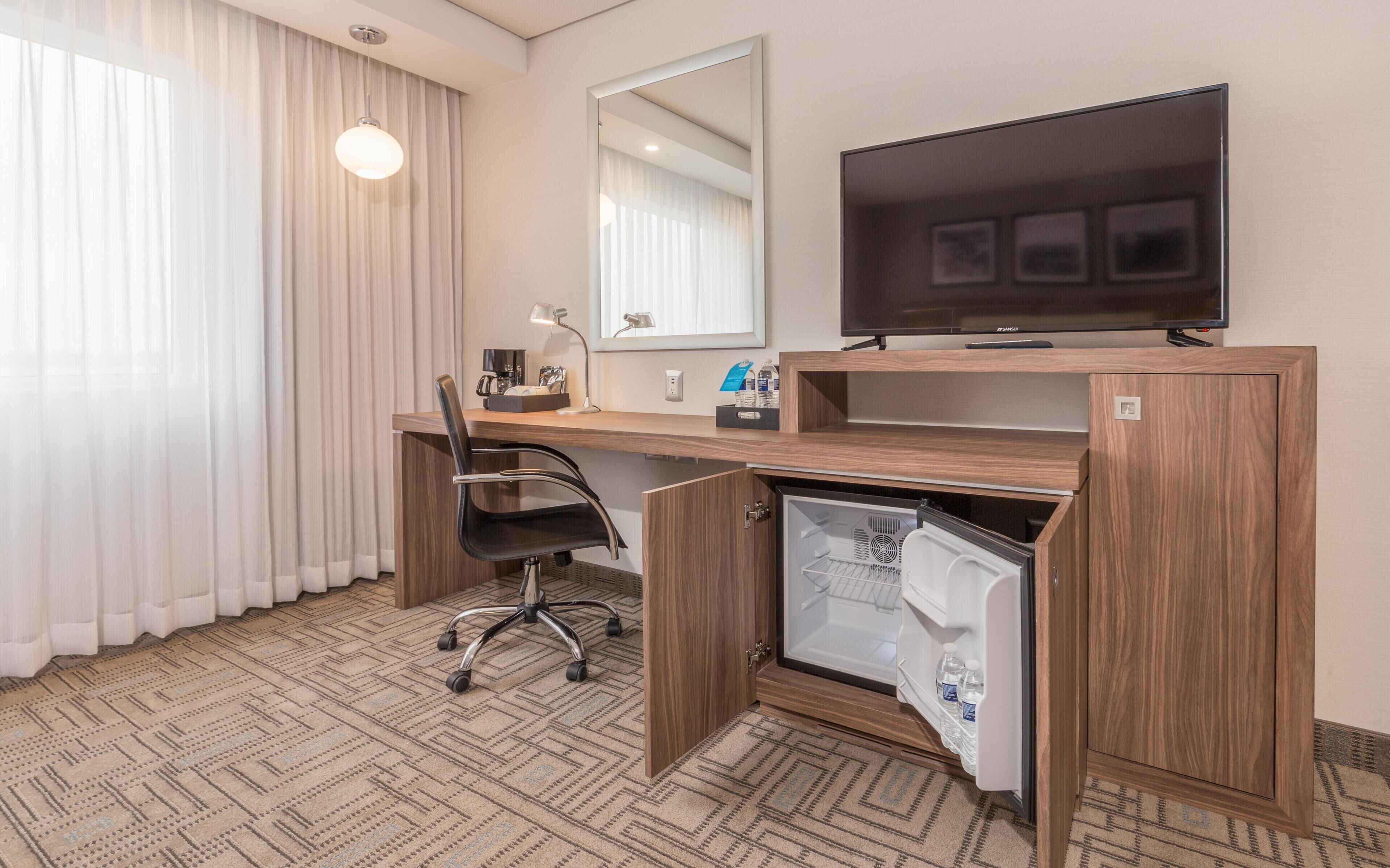 A hotel room with a wooden desk, a chair, a television, a mini-fridge, and a large window with curtains.
