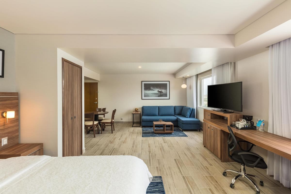 A hotel room with a bed, a desk with a computer, a TV, a sofa, and a dining area with chairs and a table.