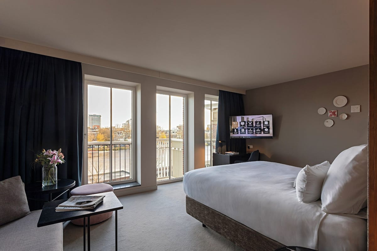 A modern hotel room with a large bed, a TV mounted on the wall, and a view of the city through the windows.