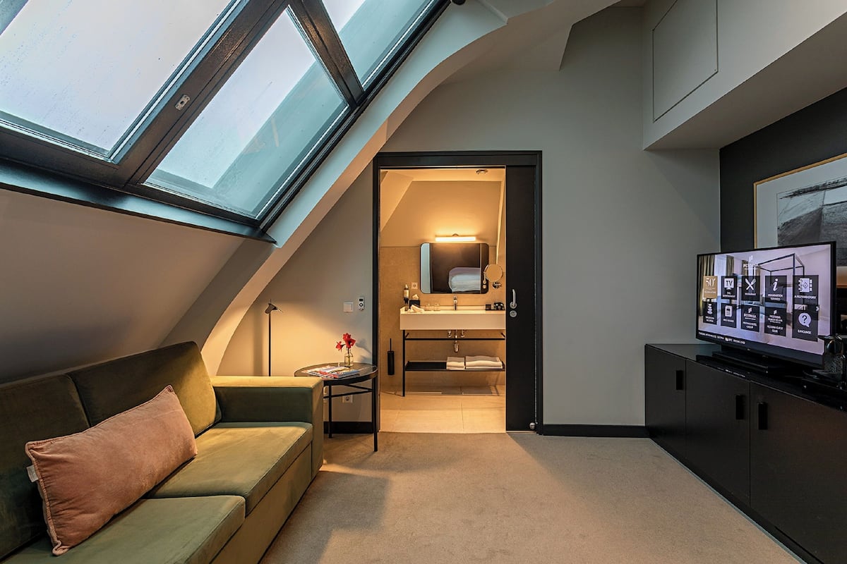 A modern hotel room with a skylight, a green sofa, a small table with a vase, a TV, and a bathroom visible through an open door.