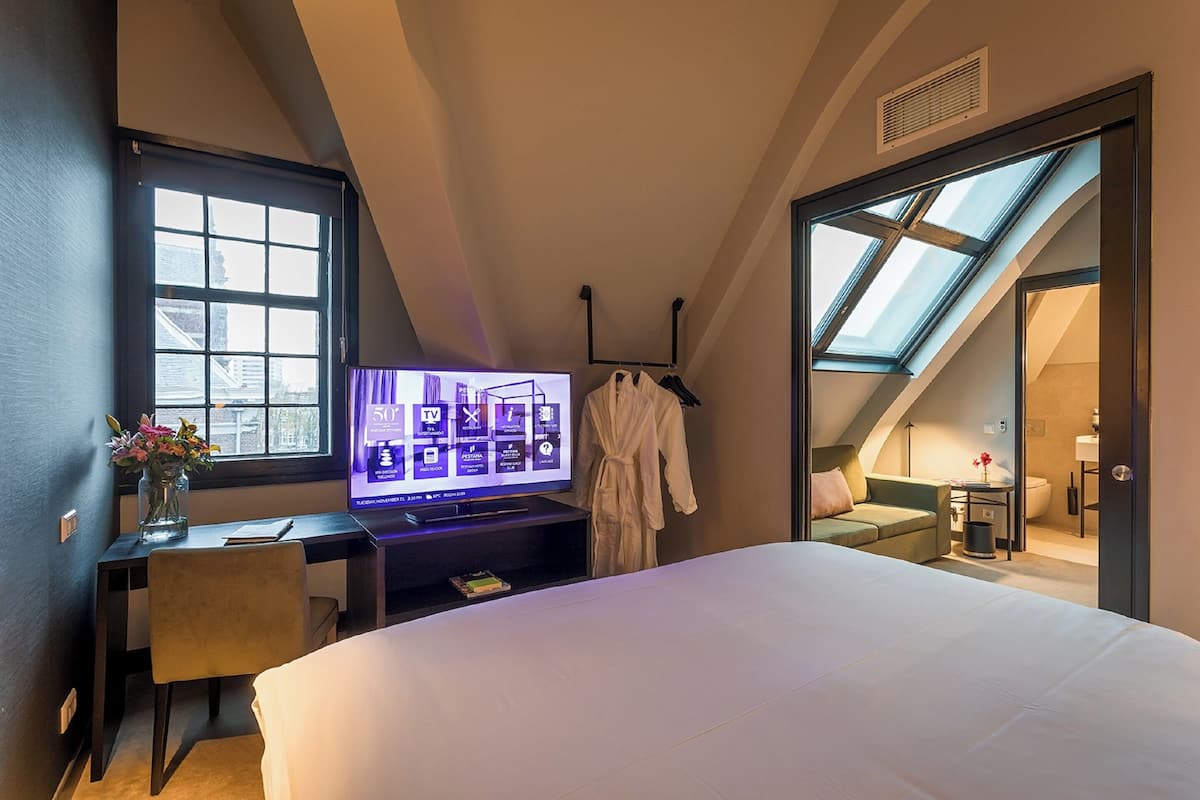 A hotel room with a large bed, a desk with a flat-screen TV, a window, and a skylight.