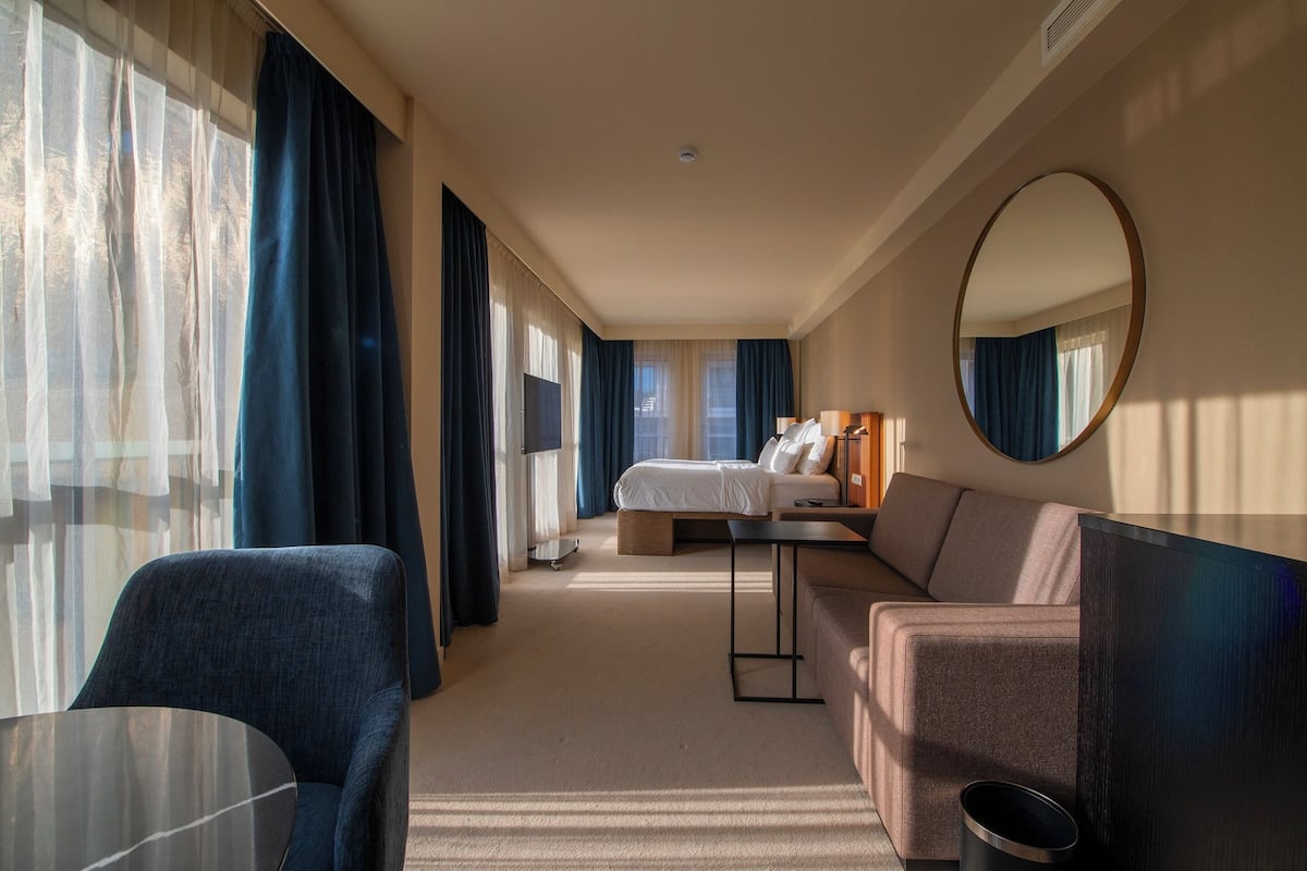 A hotel room with a bed, a sofa, a table, a mirror, and a view of the outdoors through the window.