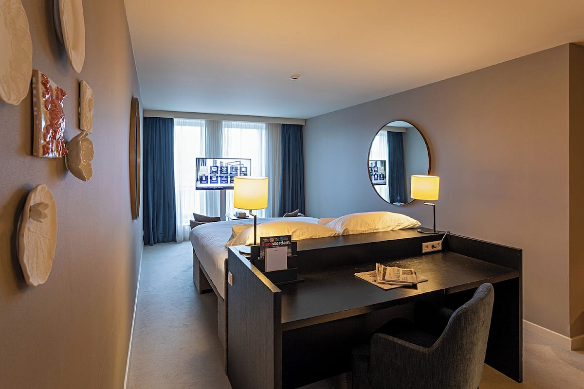 A hotel room with a bed, a desk, a chair, a round mirror, and a wall with decorative items.