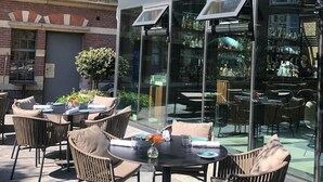 An outdoor seating area with wicker chairs and tables, a glass enclosure with a view of the interior, and a brick building in the background. - Pestana Amsterdam Riverside (Amsterdam)