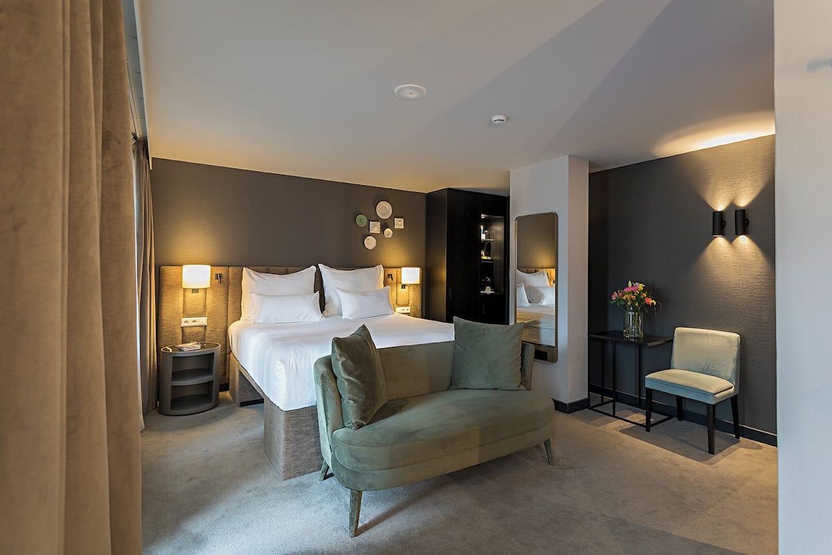 A hotel room with a large bed, bedside lamps, a sofa, a chair, and a small table with a vase of flowers.
