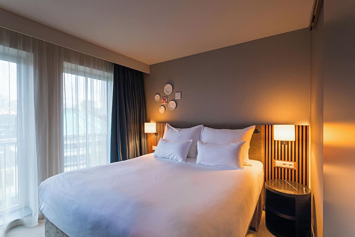 A hotel room with a large bed, two bedside lamps, a window with curtains, and a wall with framed pictures.