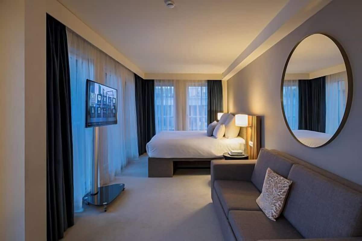 A hotel room with a bed, a sofa, a TV, a mirror, and a window with curtains.